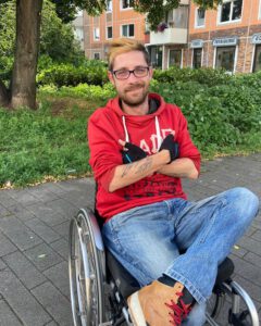 Chris sitzt in seinem Rollstuhl in einem Park. Er verschränkt selbstbewusst die Arme vor der Brust und schlägt sein linkes Bein über sein rechtes Bein. Chris trägt ein rotes Oberteil und eine blaue Jeans. Er lächelt in die Kamera.