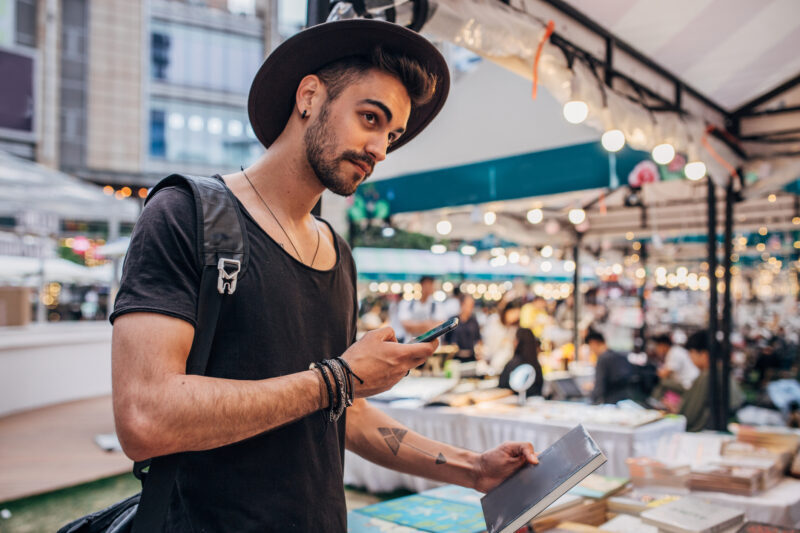 Handsome modern man with hat and beard, standing on a book fair, holding book and text messaging.