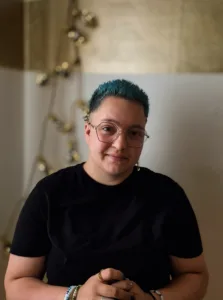 Porträt von Jo. Jo ist eine Person mit weißer Haut und kurzen, blaugefärbten Haaren. Jo trägt eine runde Brille und ein schwarzes T-Shirt. Jo lächelt in die Kamera.