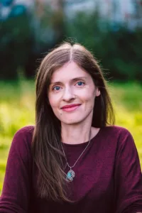 Porträt von Adina, ener Frau mitte 30 mit weißer Haut und langen hellbraunen Haaren. Sie trägt ein brombeer-farbenesOberteil und sitzt draußen.