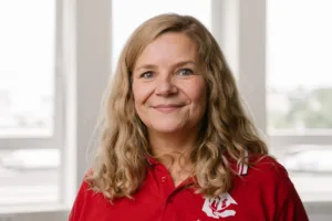 Ein Porträt von einer weißen Frau, die in die Kamera lächelt. Die hat schulterlange blonde wellige Haare und trägt ein rotes T-Shirt .