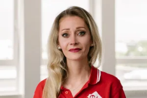 Ein Porträt von einer weißen Frau, die in die Kamera lächelt. Die hat lange blonde wellige Haare, trägt ein rotes T-Shirt und roten Lippenstift.