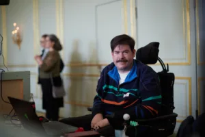 Foto von Benjamin, einem weißen Mann mit schwarzen Haaren und einem Schwarzen Schnurrbart. Er trägt ein Schwarze Jacek mit Farbenfrohen Streifen. Er sitzt in einem E-Rollstuhl.