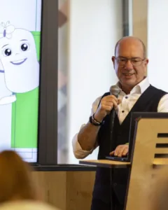 Foto von Christian, einem weißen Mann mit einer Brille und ohne Haare auf dem Kopf. Er trägt ein weißes Hemd und eine schwarze Weste undi spricht auf dem Foto in ein Mikrofon.