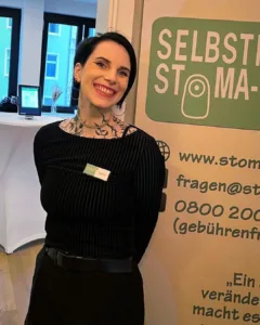 Foto von Jana, einer Frau Ende 20 mit weißer Haut, vielen Tattoos, kurzen schwarzen Haaren und schwarzer Kleidung. Sie lacht