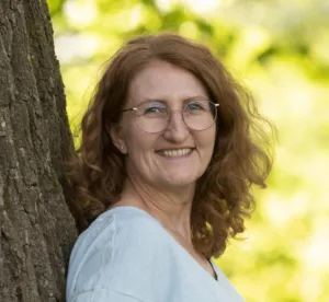 Porträtfoto von Karin, einer weißen Frau mit rötlich-blonden langen Locken. Sie trägt eine Brille und ein weißes Oberteil. Sie lehnt an einem Baum.