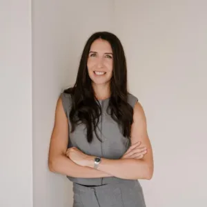 Ein Porträt von Anja. Anja ist eine weiße Frau mit langen schwarzen Haaren. Sie trägt ein graues Kleid und lehnt an einer Weißen Wand. Sie lächelt und verkreuzt die Arme selbstbeswusst