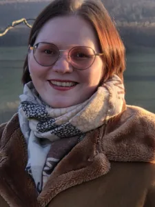 Andrea ist eine weiße Frau mit schulterlangen rot-brauen Haaren. Sie strahlt in die Kamera und trägt eine braune Jacke, einen schal und eine braun-getönte Brille.