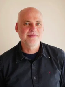 Weißer Mann, mittleren Alters mit Halbglatze un weißem Bart trägt ein schwarzes Hemd und steht vor einer weißen Wand.